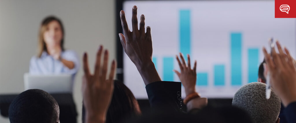 A presenter stands in front of a screen displaying a bar graph on a PowerPoint slide, while audience members raise their hands, indicating participation or questions. The background is slightly blurred, focusing on the audience's hands in the foreground.