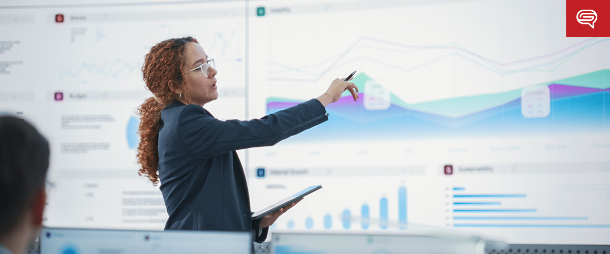 A person with curly hair and glasses, wearing a suit, is presenting data displayed on large screens. They are pointing at charts and graphs with a stylus in one hand and holding a tablet that resembles an advanced digital flipbook in the other. The background shows various financial or statistical data.