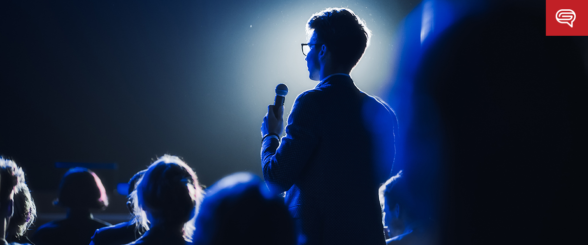 A person is seen from behind, standing in front of an audience and speaking into a microphone. The setting is dimly lit with a spotlight illuminating the speaker, reminiscent of frames in a flipbook. Several audience members are visible in the foreground with their faces obscured.