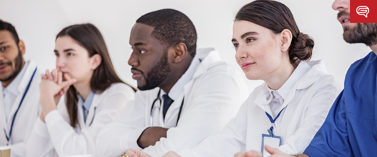 A group of five attentive medical professionals in white lab coats sits at a table, likely engaged in a life science presentation. They pay close attention to something out of frame, badges indicating their roles in the medical field.