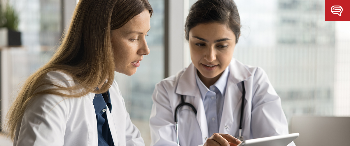 Two female doctors in white lab coats are sitting at a table, discussing information displayed on a tablet like flipping through a digital flipbook. One doctor is pointing at the tablet while the other listens attentively. In the background, large windows reveal a bright, blurred cityscape.