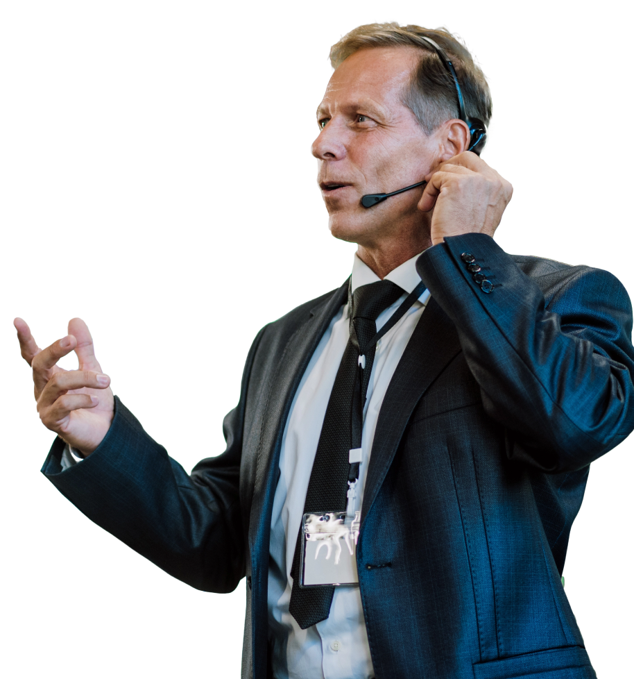 A person in a suit and tie is speaking while wearing a headset microphone, gesturing with one hand and appearing to give a speech. They have a lanyard with an ID card around their neck, embodying the confidence of sales presentations experts.