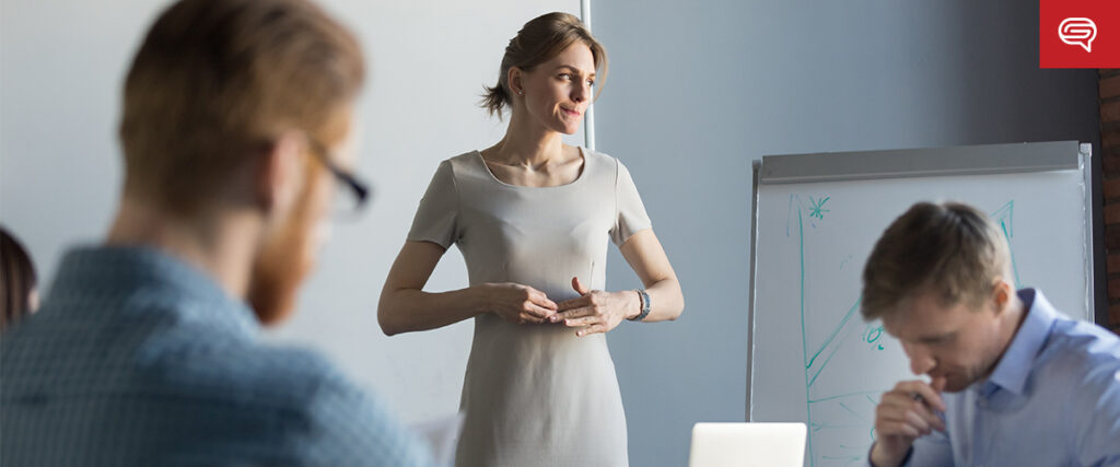 A woman is standing and speaking in front of a group, gesturing with her hands. Two men are sitting and listening attentively, one with his hand near his mouth. A whiteboard with diagrams is in the background, possibly showcasing animations in pitch decks, and a small red logo is visible in the top right corner.