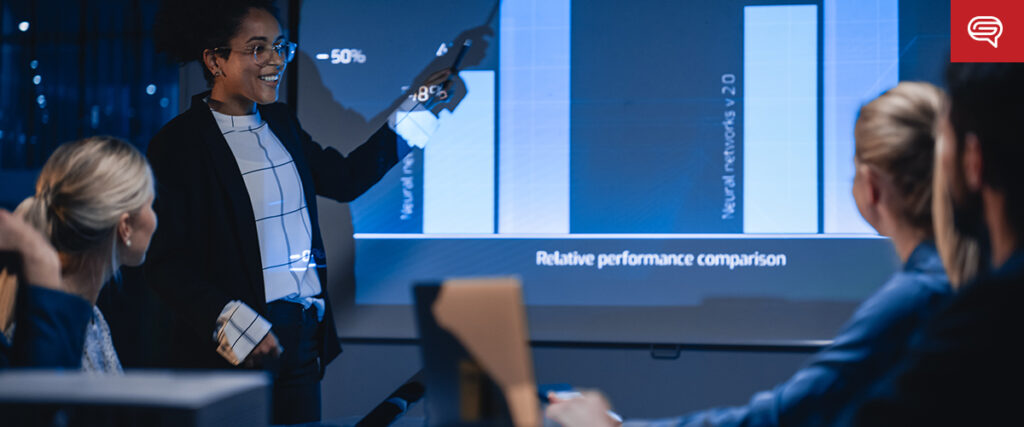 A person is presenting a quality earnings report displayed on a large screen to four seated colleagues in a dimly lit room. The screen shows bar charts labeled "Relative Performance Comparison" and mentions "Neural Networks." The presenter points at the chart with a smile.