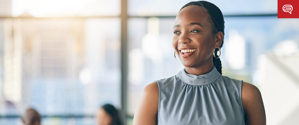 A smiling woman stands in a brightly lit office with several people working in the background. She is dressed in a sleeveless, high-collared top and has her hair tied back. Large windows provide a view of a cityscape outside, hinting at her recent success with quality earnings report presentations.