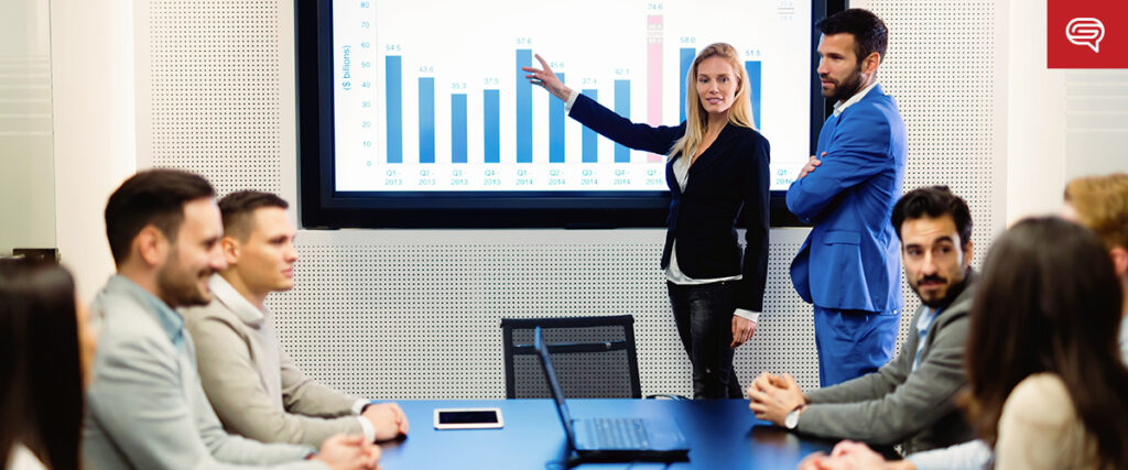 A woman stands at the front of a conference room, pointing at a bar graph on a screen. A man in a blue suit stands beside her. Seven people sit around a table listening attentively as animations in pitch decks bring data to life. A laptop is open on the table, and the background shows a modern office setting.