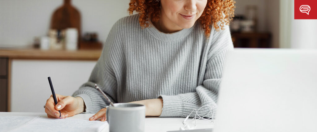 A person with curly hair is sitting at a table, writing in a notebook with one hand and using a laptop with the other, likely working on animations in pitch decks. A gray mug and a pair of white earbuds are placed on the table. The background is blurred, suggesting an indoor setting.