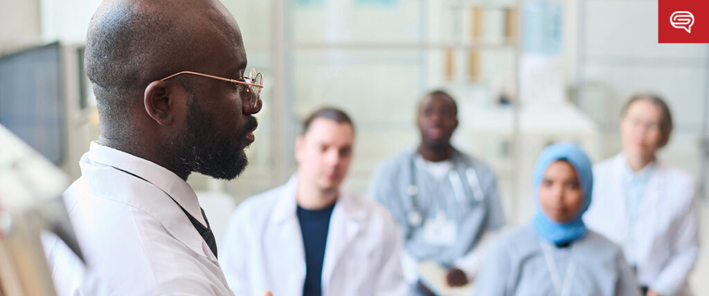 A person in a white coat and glasses is presenting to a group, also in lab coats, seated in a bright room. The attentive group suggests this lecture uses case study slides to influence decisions in the medical or scientific setting.