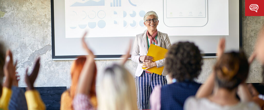 A person with short gray hair and glasses stands smiling in front of a projected presentation, likely a pitch deck. They are holding a yellow folder. An audience is in the foreground, some with raised hands, indicating an engaging seminar or classroom atmosphere.