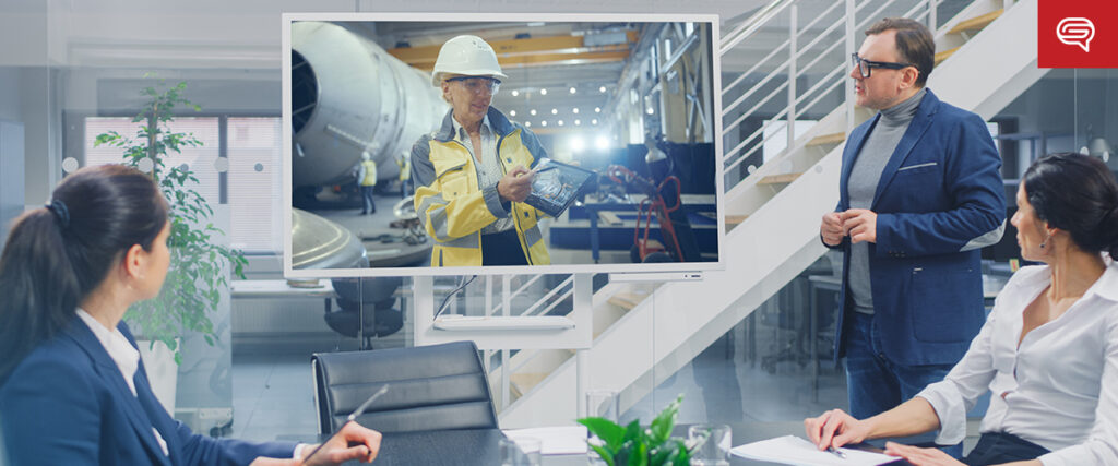 People in a modern office meeting room watch a seed funding round presentation on a large screen. The screen displays an industrial worker in a hard hat discussing a piece of equipment. The room features a glass wall, stairs, and a potted plant.