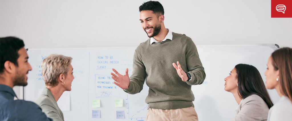 A man stands smiling, engaging with four seated colleagues during a seed funding round presentation. They are gathered around a table, with a whiteboard full of notes and graphs in the background. The atmosphere appears collaborative and positive.