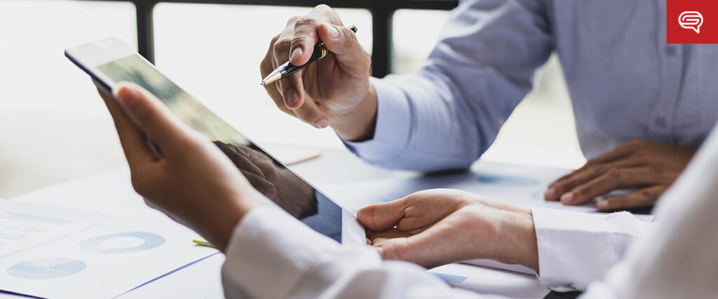 Two people discuss work at a desk, one pointing a pen at a tablet screen, amidst documents with charts and graphs. The focus is on collaboration and data analysis as they prepare for seed funding round presentations.