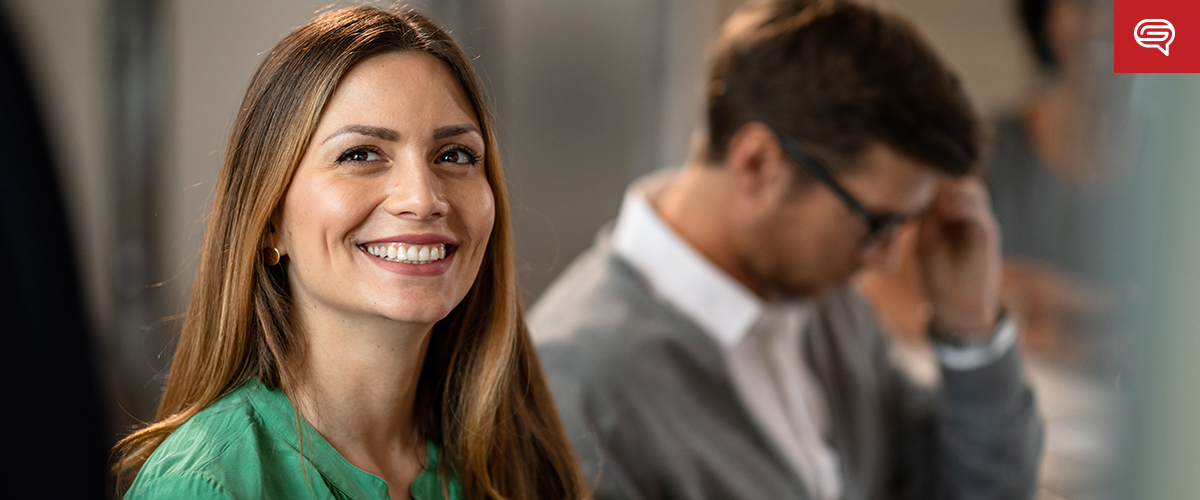 A woman with long brown hair smiles while wearing a green blouse, embodying the ideal client experience. She sits in an office setting, with a man in the background adjusting his glasses. The atmosphere is professional and warm, akin to a polished slide for consulting firms.