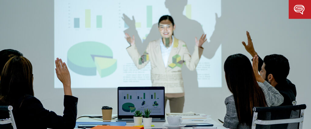 A woman gives a presentation in a conference room, gesturing towards charts on the client experience slide projected on a screen. Four colleagues sit at the table with a laptop, coffee cup, and notebook. One person raises a hand, appearing engaged—a typical scene for consulting firms.