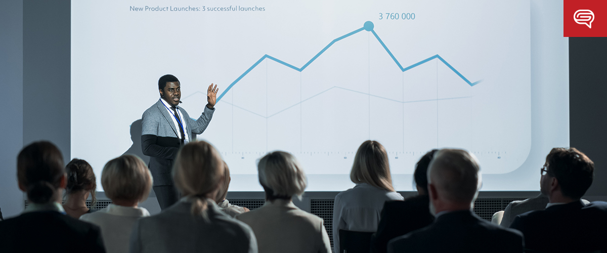 A presenter in a suit gestures towards a client experience slide featuring a projected line graph showing product launch data. The graph highlights peaks with the label "3,760,000". Attendees from consulting firms are seated in rows, attentively facing the screen.