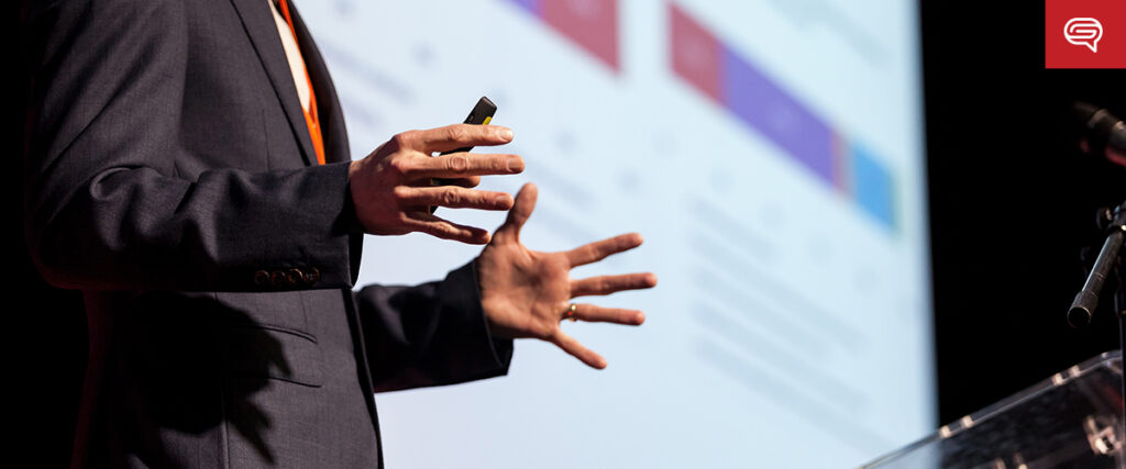 A person in a suit gestures expressively with their hands while standing in front of a presentation screen displaying charts and text, enhancing the client experience. A microphone is visible to the right, emphasizing their dynamic presentation style frequently seen in consulting firms.